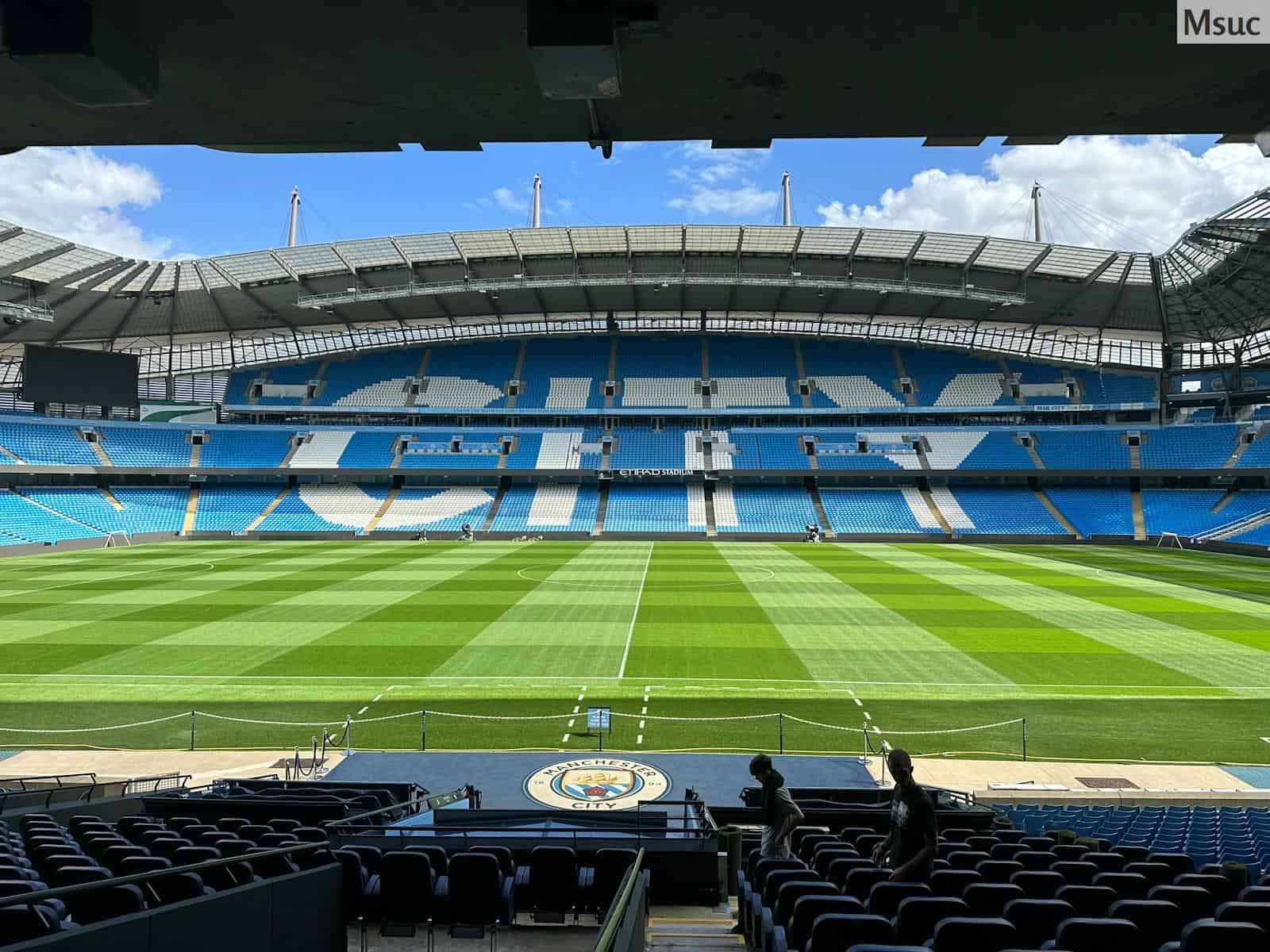 Panoramic view of Etihad Stadium, showcasing the empty football pitch and stands under a clear blue sky.